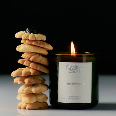 Candle labeled 'Flamingo Estate' with a stack of cookies on a reflective surface.