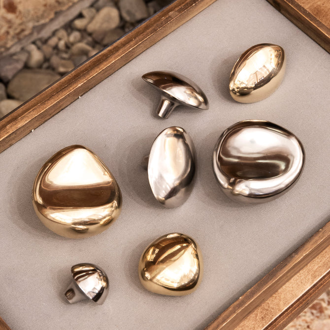Seven polished metallic pebble-shaped hardware displayed on a light gray velvet-lined jewelry box, with a terrazzo-patterned surface visible in the background.